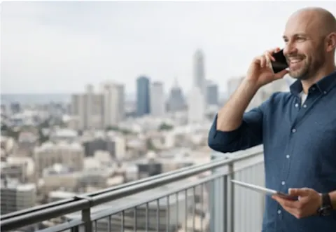 Homme expatrié au téléphone sur un balcon avec vue urbaine, représentant un accompagnement en chasse immobilière