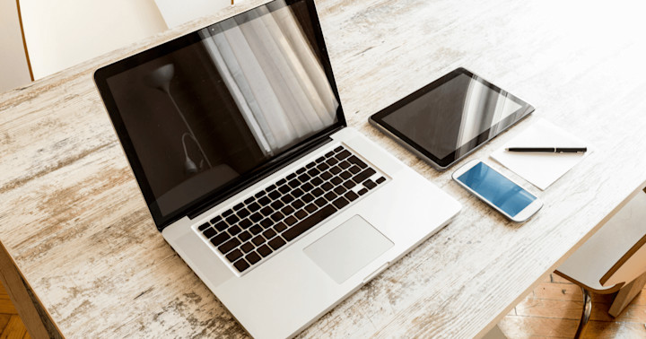 A laptop, tablet, smart phone, and a pad of paper with a pen sitting on a wooden table.