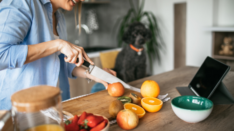 Woman cuts fruit in kitchen while dog watches in background