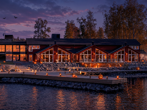 En byggnad vid sjön i skymningen med stora fönster och halloween-lyktor placerade längs bryggan och uteplatsen, med höstträd och en färgsprakande himmel i bakgrunden.