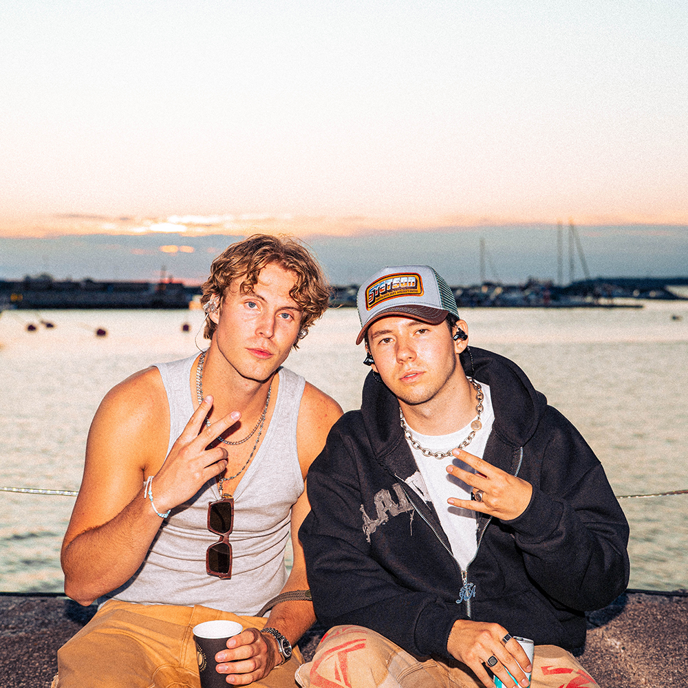 Två unga män sitter vid vattnet i solnedgången, den ene i linne och med en kopp i handen, den andre i keps och huvtröja; båda gör handgester.