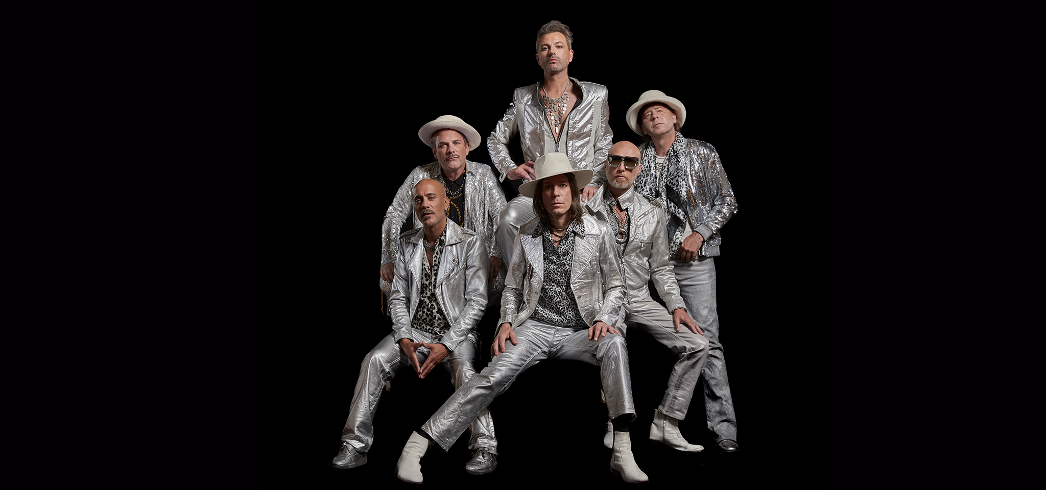 Sex män poserar mot en svart bakgrund, alla klädda i matchande silverglänsande kostymer, hattar och mönstrade skjortor, med allvarliga ansiktsuttryck.