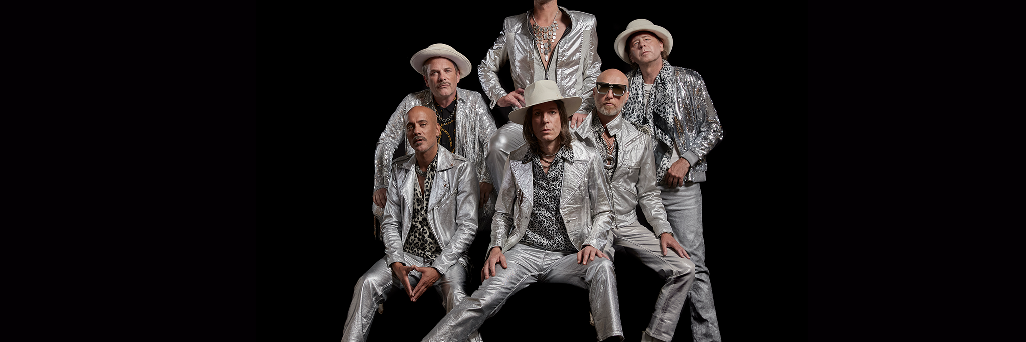 Sex män poserar mot en svart bakgrund, alla klädda i matchande silverglänsande kostymer, hattar och mönstrade skjortor, med allvarliga ansiktsuttryck.