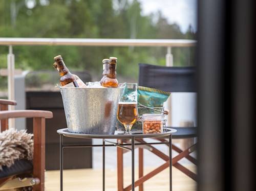 En metallhink med ölflaskor i is, ett glas öl, snacks och en stol på en uteservering med räcke och träd i bakgrunden.
