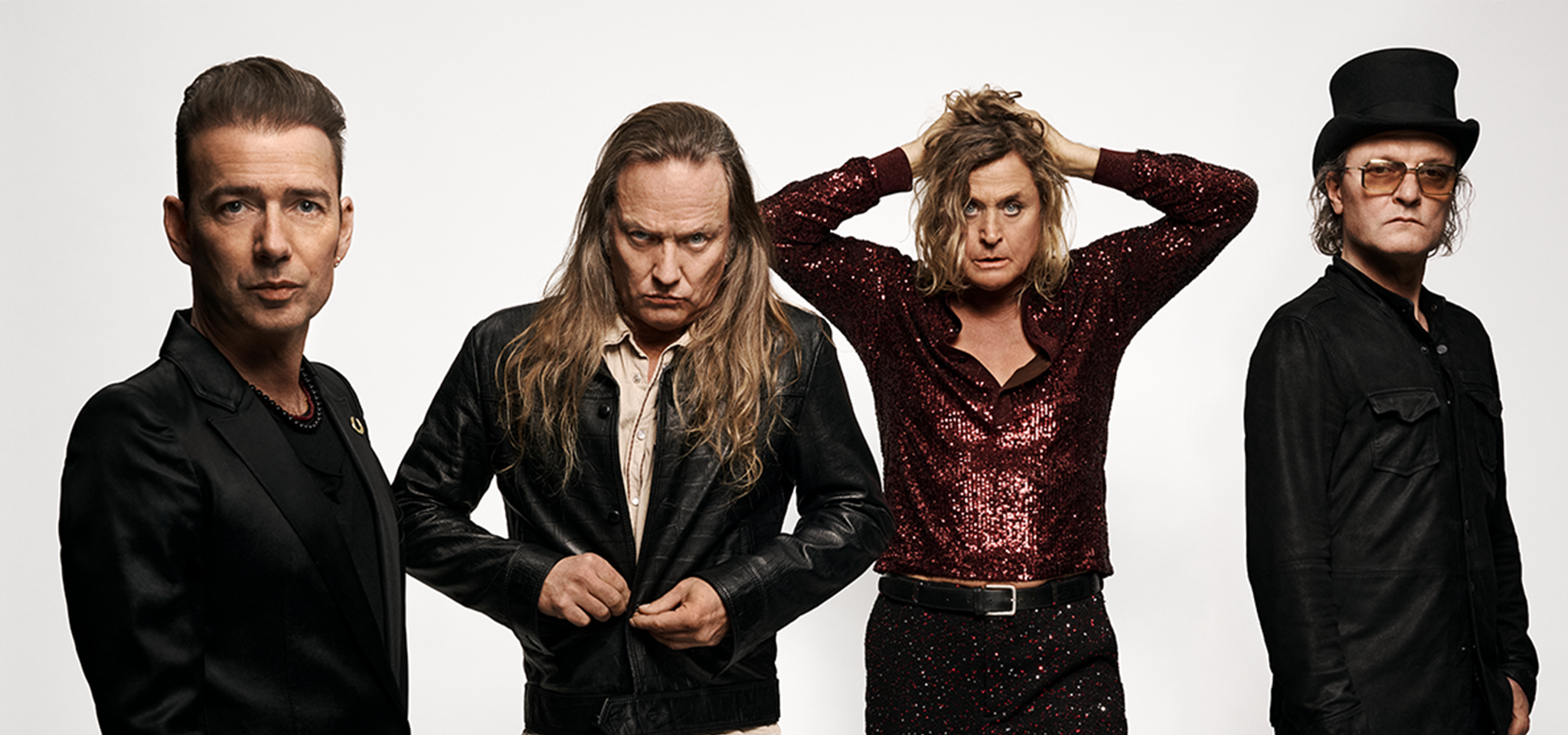 Fyra män står mot en vit bakgrund, alla med olika ansiktsuttryck och klädstilar, bland annat läderjackor, en paljettskjorta och en hög hatt.
