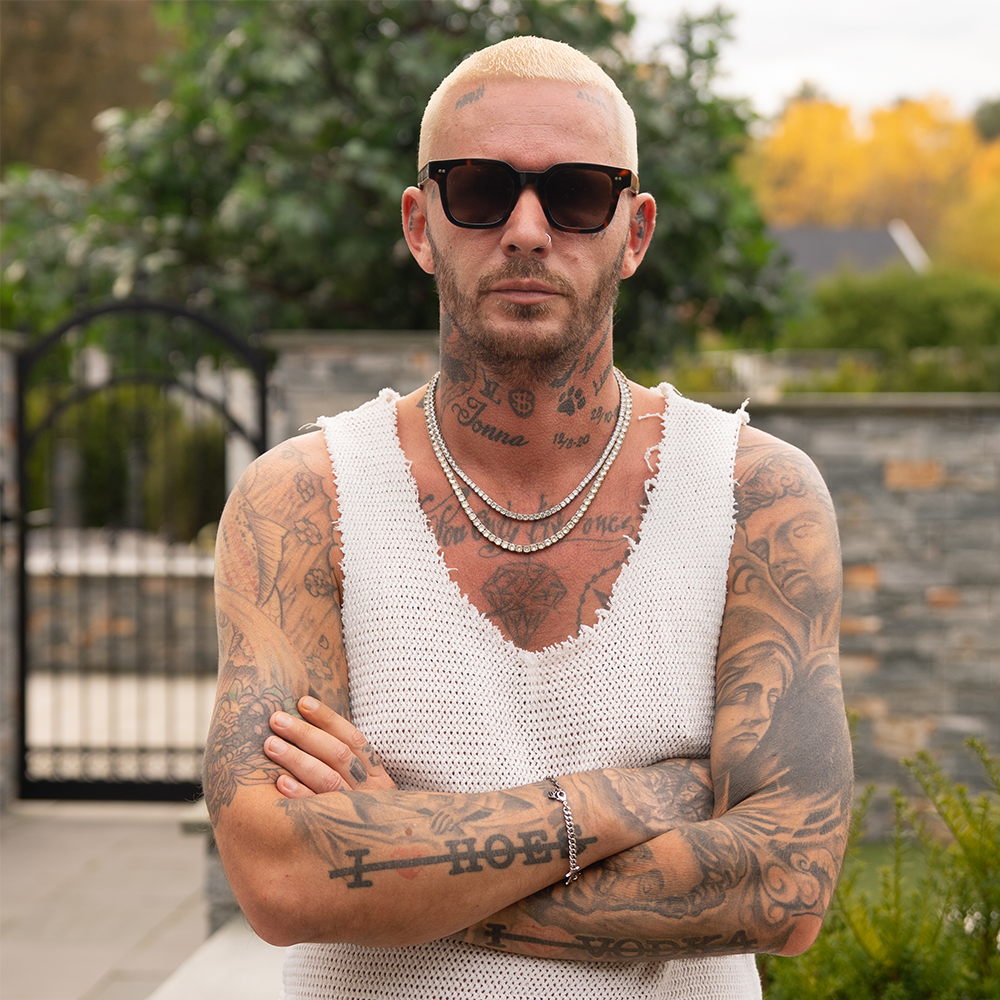 A man with bleached hair, sunglasses, and extensive tattoos stands outdoors with arms crossed, wearing a sleeveless white knit top and layered necklaces.