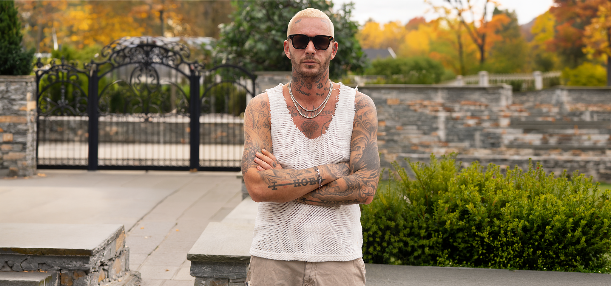 A tattooed man with shaved blond hair, sunglasses, and a sleeveless shirt stands outdoors with arms crossed, in front of a stone fence and garden.