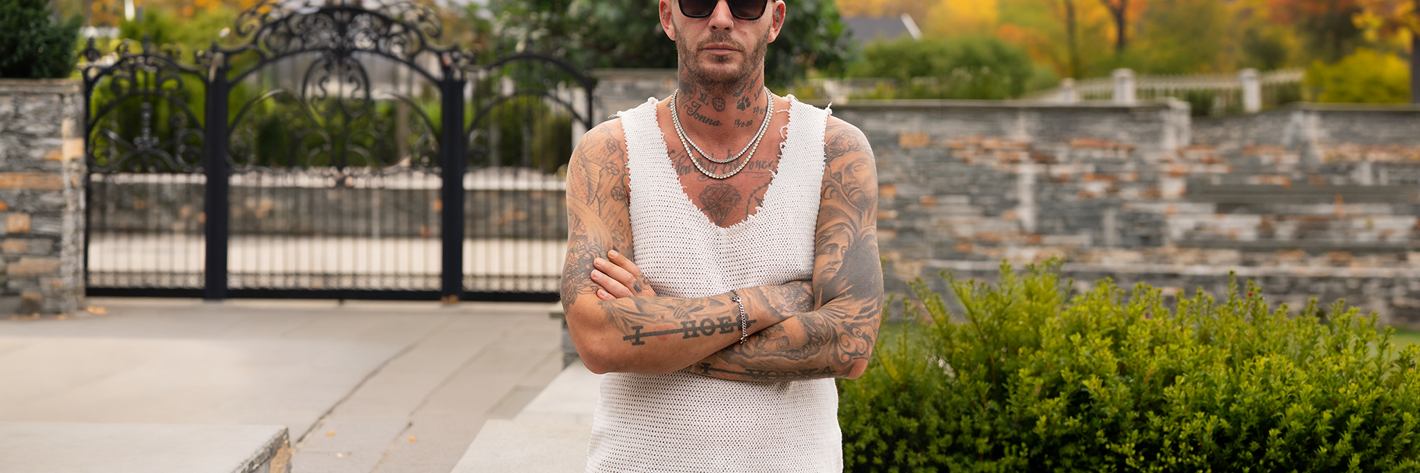 A tattooed man with shaved blond hair, sunglasses, and a sleeveless shirt stands outdoors with arms crossed, in front of a stone fence and garden.