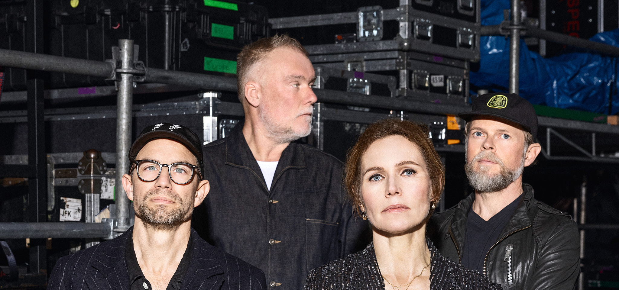 Fyra vuxna står framför staplade lådor och utrustning i vad som ser ut att vara ett backstageområde. Tre är vända framåt, en tittar åt sidan.