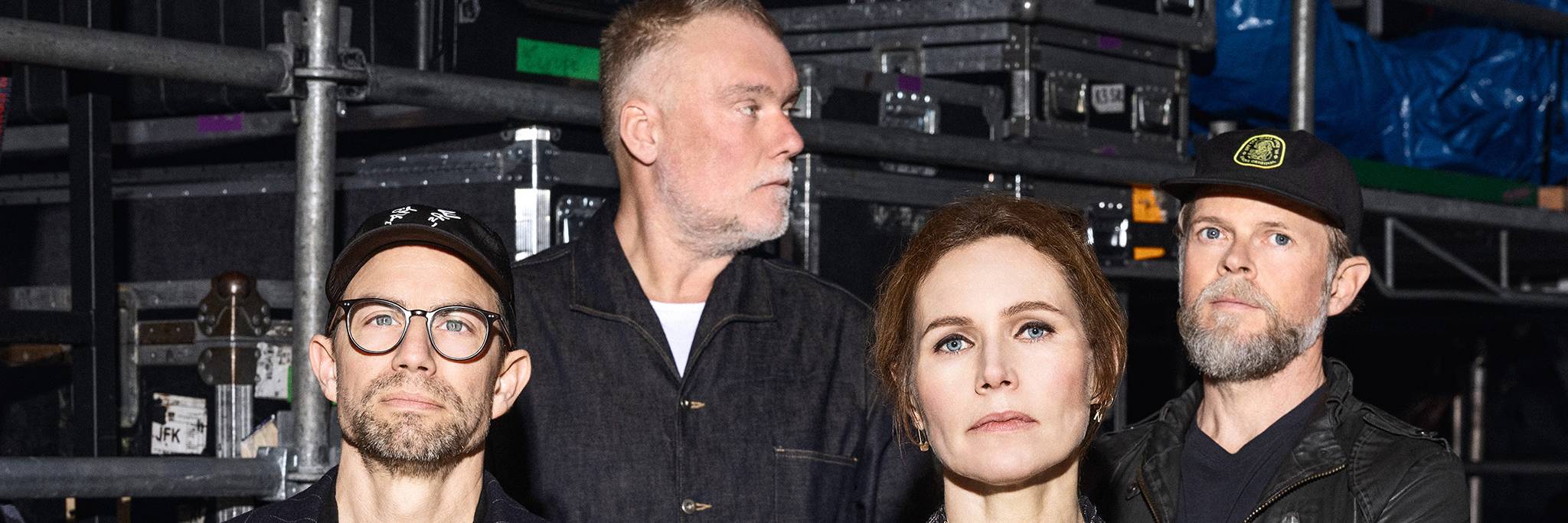 Fyra vuxna står framför staplade lådor och utrustning i vad som ser ut att vara ett backstageområde. Tre är vända framåt, en tittar åt sidan.