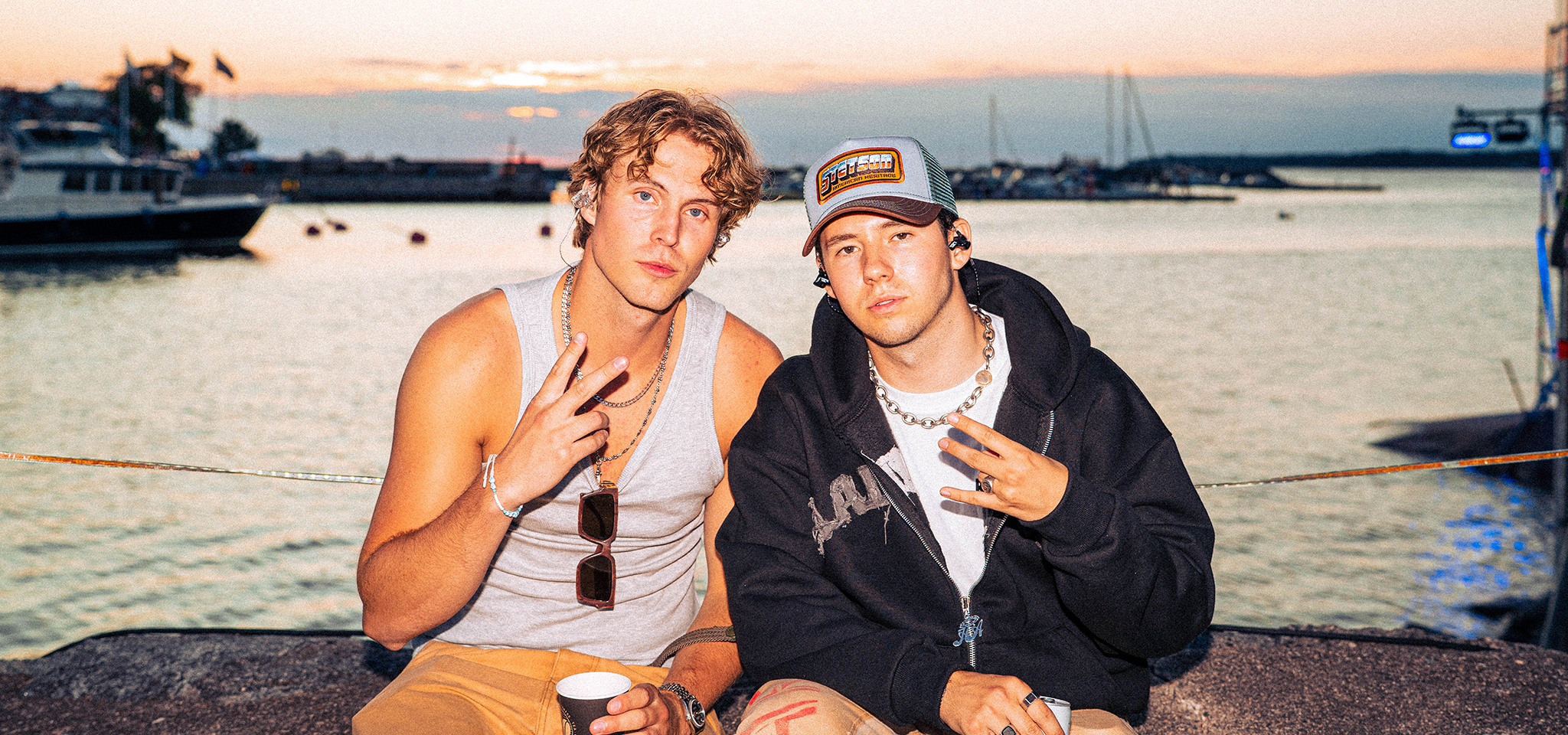 Två unga män sitter vid vattnet i solnedgången och gör fredstecken med sina händer. Den ena bär ett linne och solglasögon, den andra en huvtröja och en basebollkeps.