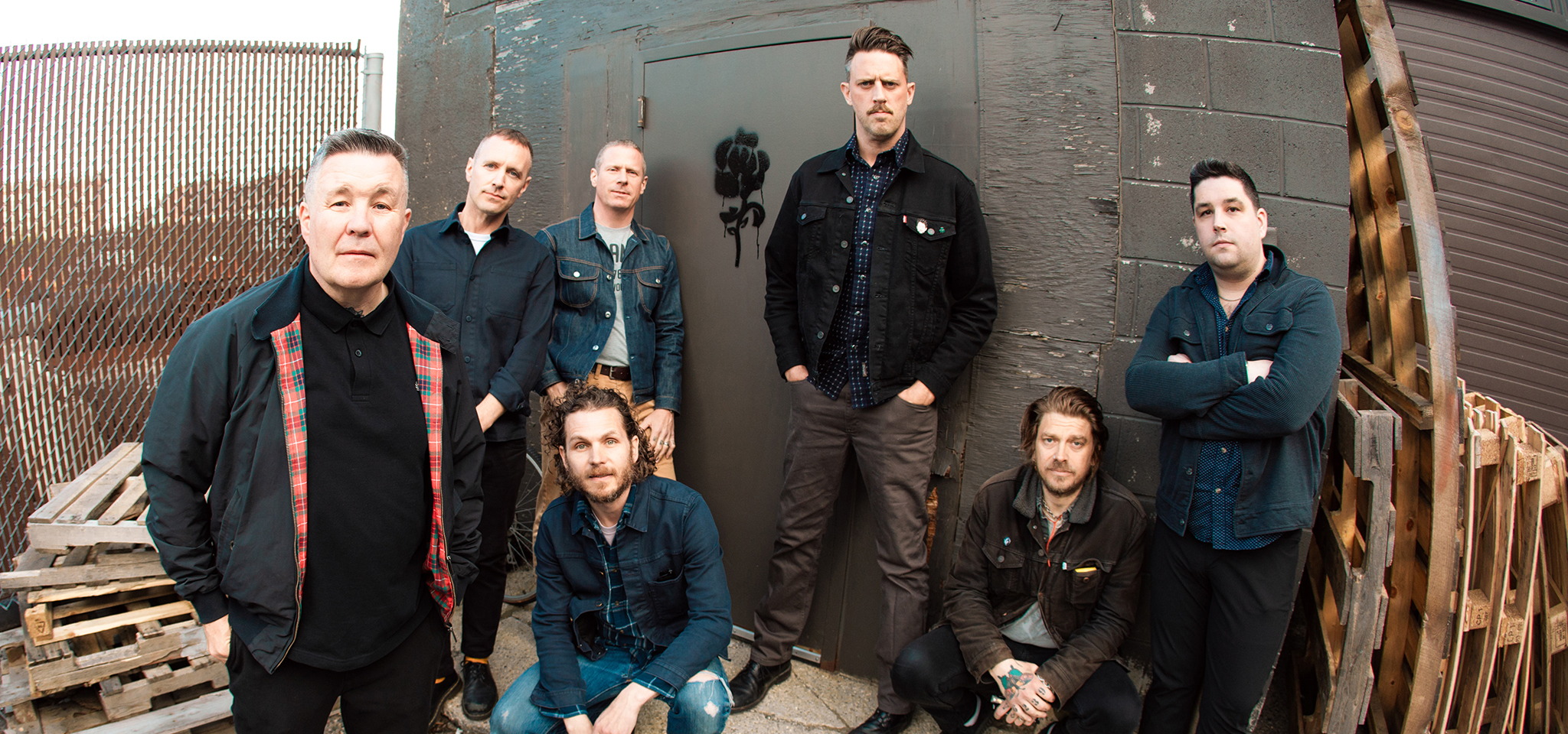 Sju män poserar för ett gruppfoto utomhus intill en betongbyggnad och träpallar, mestadels klädda i mörka fritidskläder.