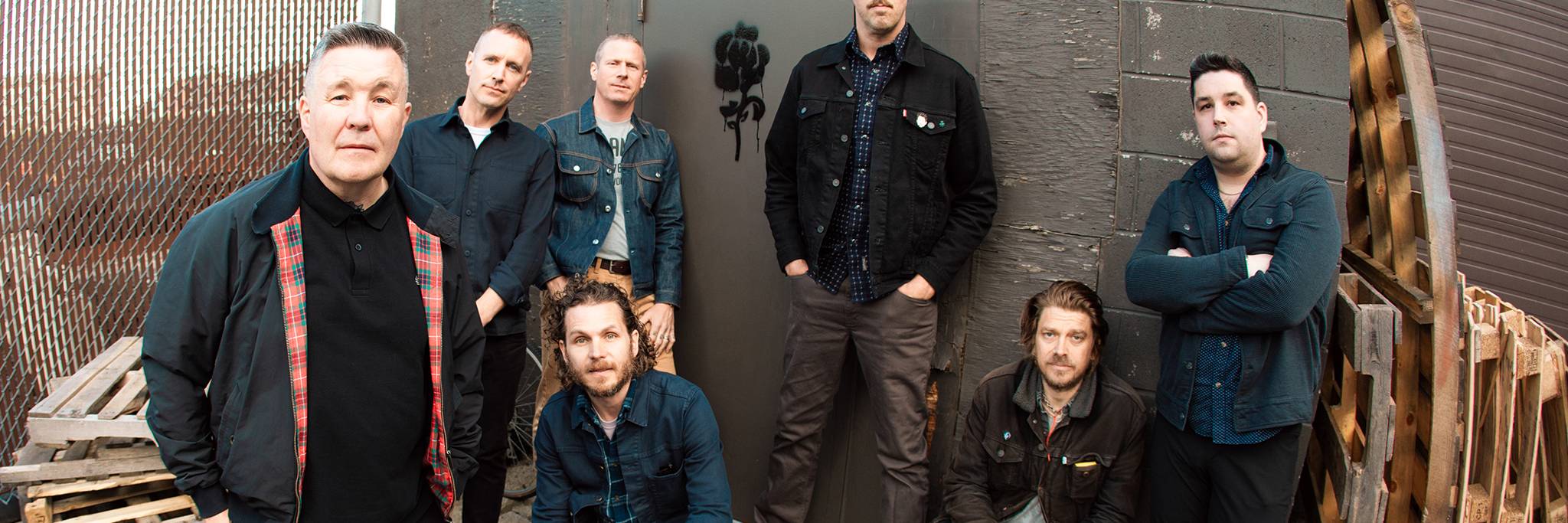 Sju män poserar för ett gruppfoto utomhus intill en betongbyggnad och träpallar, mestadels klädda i mörka fritidskläder.