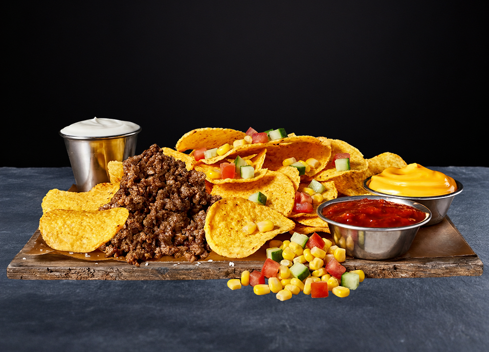 A platter of nachos topped with ground beef, diced vegetables, and served with sides of sour cream, cheese sauce, salsa, and corn on a wooden board against a black background.