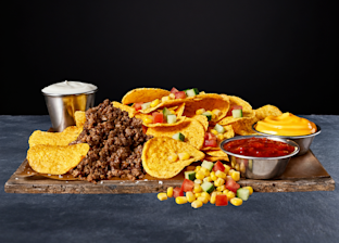 A platter of nachos topped with ground beef, diced vegetables, and served with sides of sour cream, cheese sauce, salsa, and corn on a wooden board against a black background.
