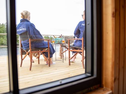 Två personer i blå kläder sitter på en träterrass vid vattnet, med ryggen vänd mot betraktaren, med drycker på ett bord, sett genom ett fönster.