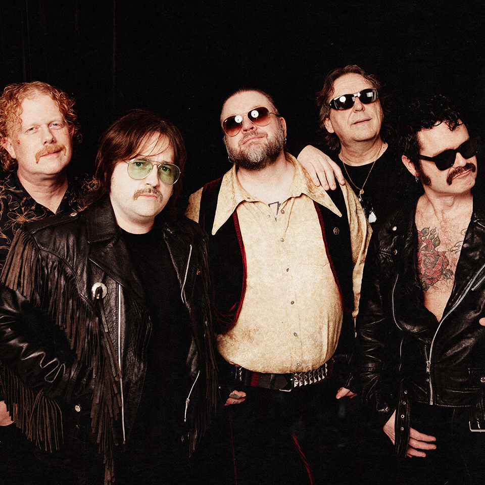 Fem män med solglasögon och kläder i vintagestil poserar tillsammans mot en mörk bakgrund.