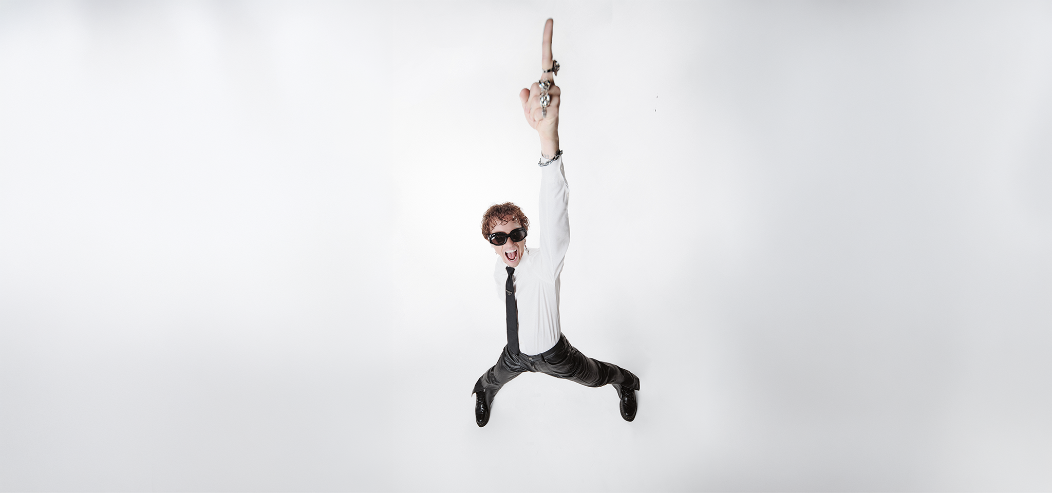 En man i vit skjorta, slips och solglasögon står med benen isär och pekar uppåt med ena armen mot en vit bakgrund.