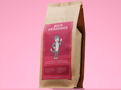 A brown bag of coffee labeled “Maja Graddnos” with a cartoon cat illustration and text indicating medium roast brew coffee, set against a pink background.