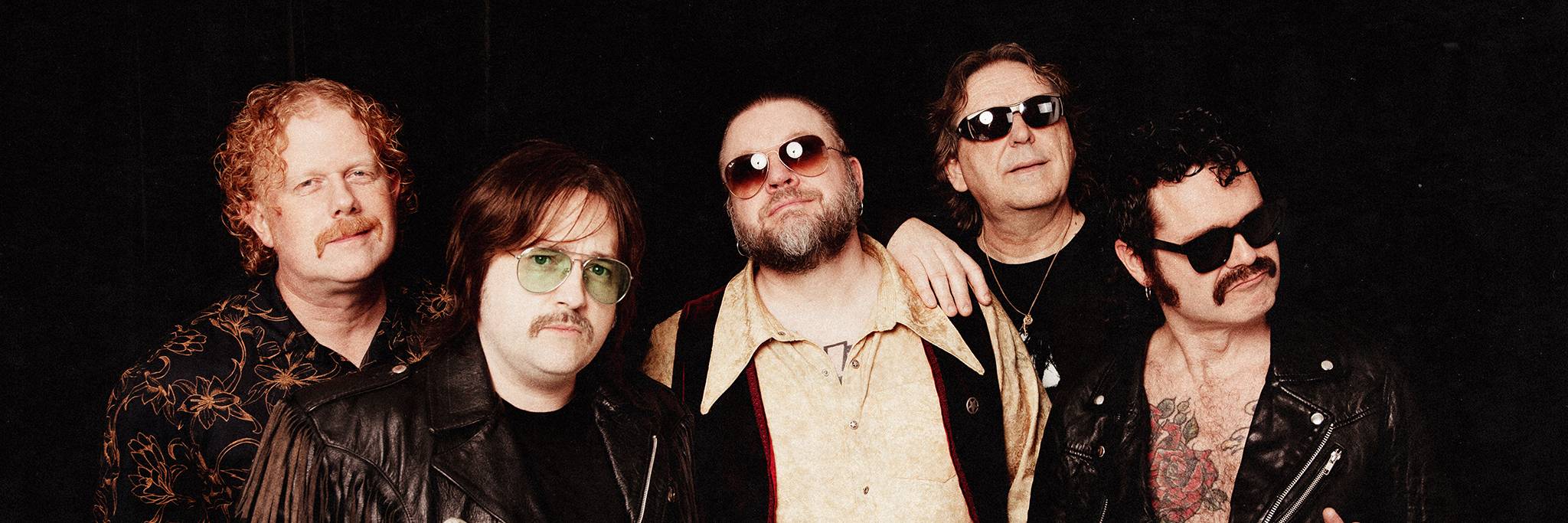 Fem män poserar tillsammans mot en mörk bakgrund, iklädda solglasögon och olika outfits, bland annat skinnjackor och mönstrade skjortor.