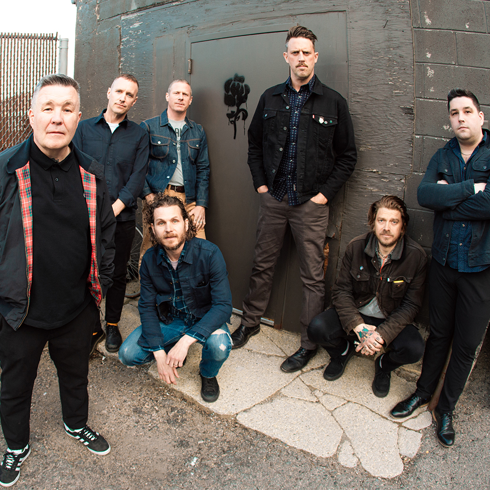 Sju män står och knäböjer framför en grå industriport och poserar för ett gruppfoto. De flesta är klädda i mörka jackor och jeans och har neutrala ansiktsuttryck.