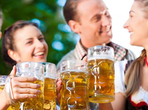Four people outdoors smiling and holding large mugs of beer, appearing to celebrate or make a toast.