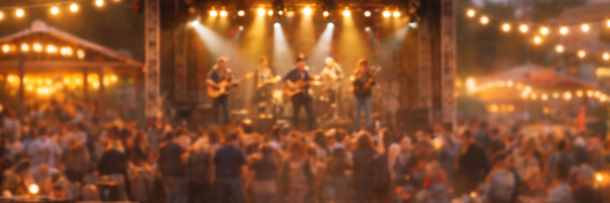En stor folkmassa samlas framför en utomhusscen där ett band uppträder under ljusslingor i skymningen.