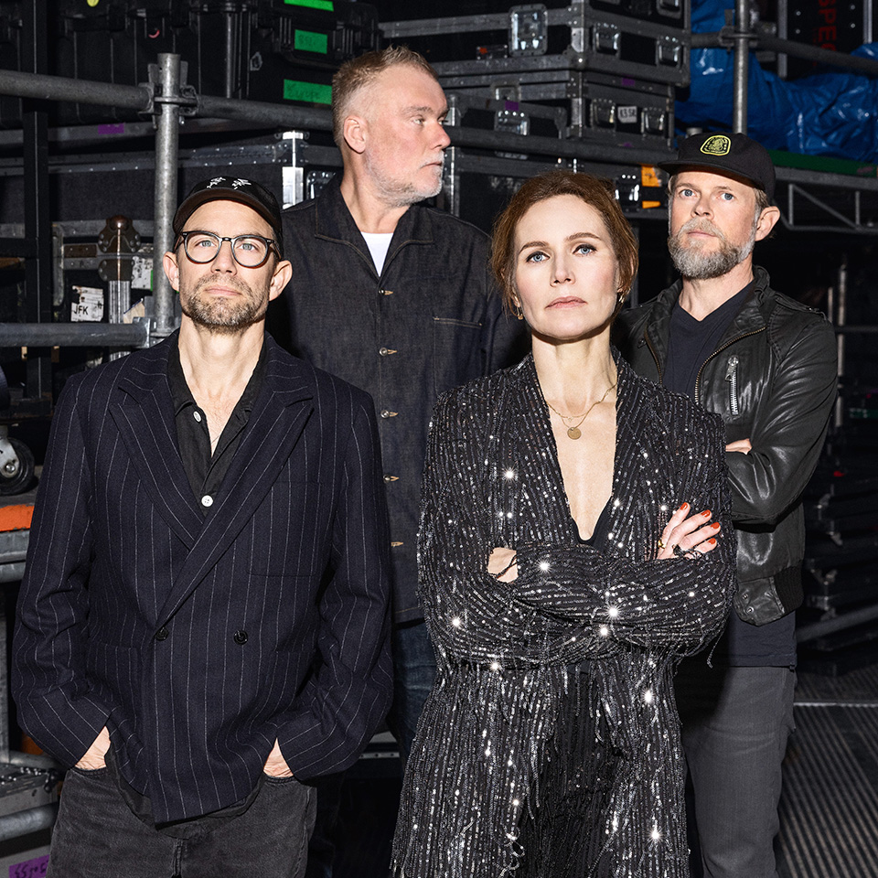 Fyra vuxna står tillsammans framför staplade utrustningslådor; tre män bär mörka jackor och hattar, medan en kvinna står i en glittrande svart outfit med armarna i kors.