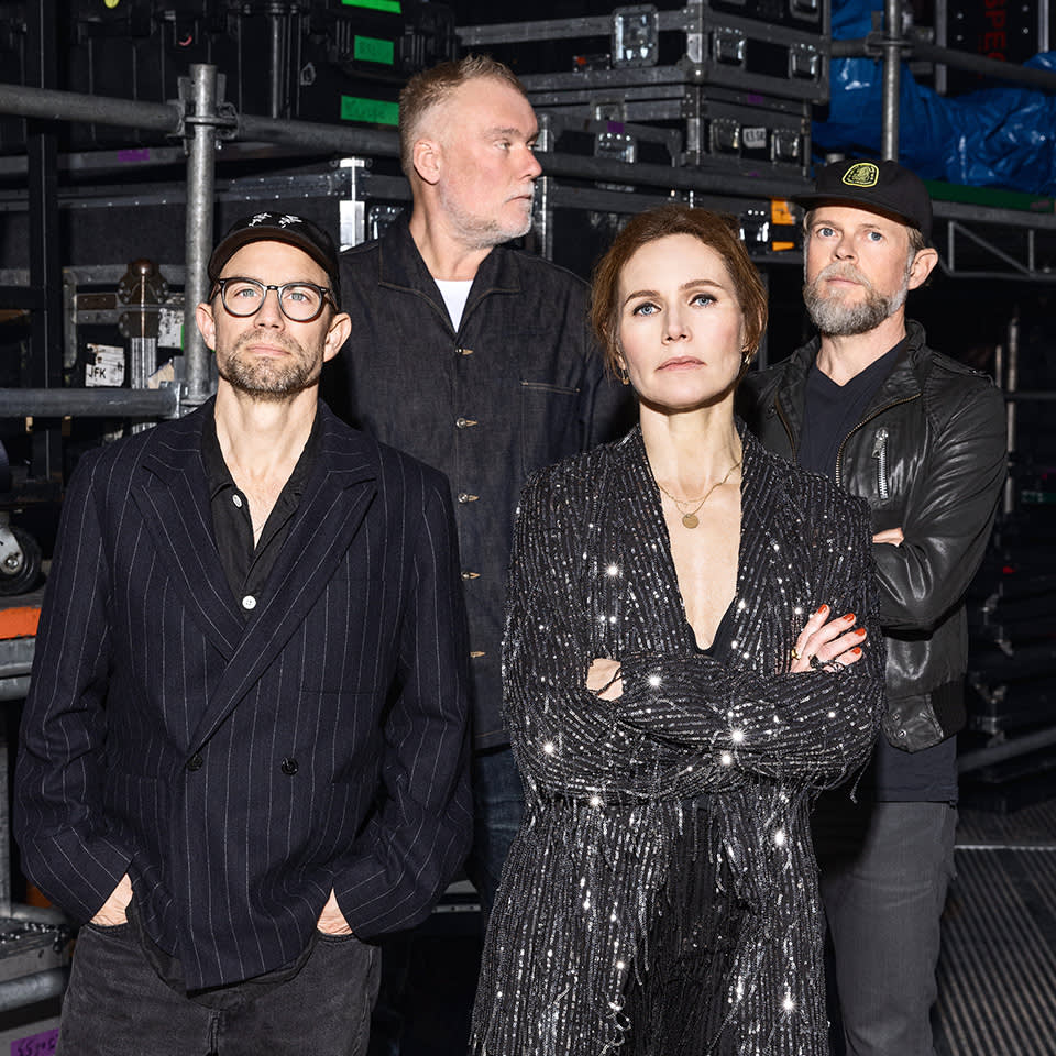 Fyra vuxna står tillsammans framför staplade utrustningslådor; tre män bär mörka jackor och hattar, medan en kvinna står i en glittrande svart outfit med armarna i kors.