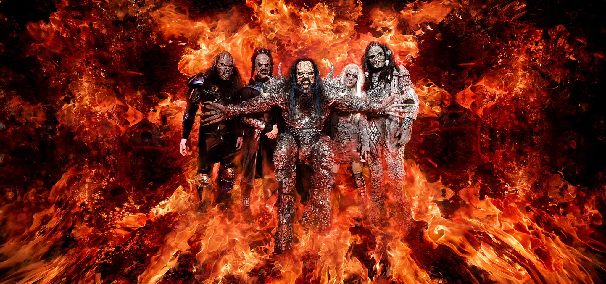 Five people in elaborate monster costumes stand together, surrounded by dramatic flames and fiery background effects.