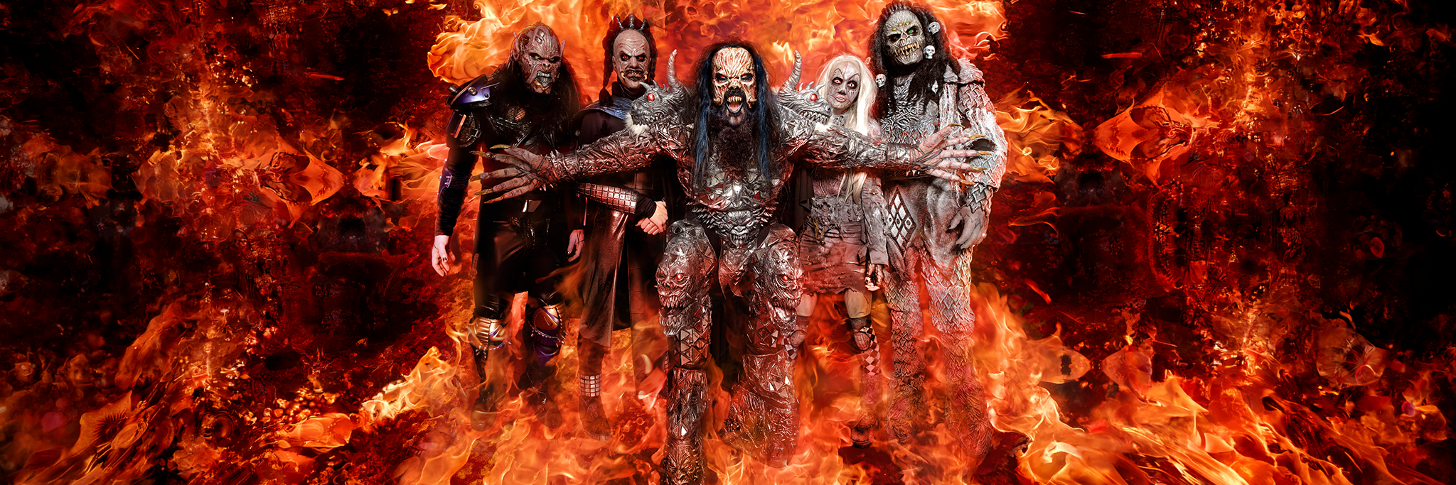 Five people in elaborate monster costumes stand together, surrounded by dramatic flames and fiery background effects.