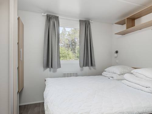 Ett litet, minimalistiskt sovrum med en prydligt bäddad säng, ett fönster med grå gardiner, en garderob och hyllor ovanför sängen.