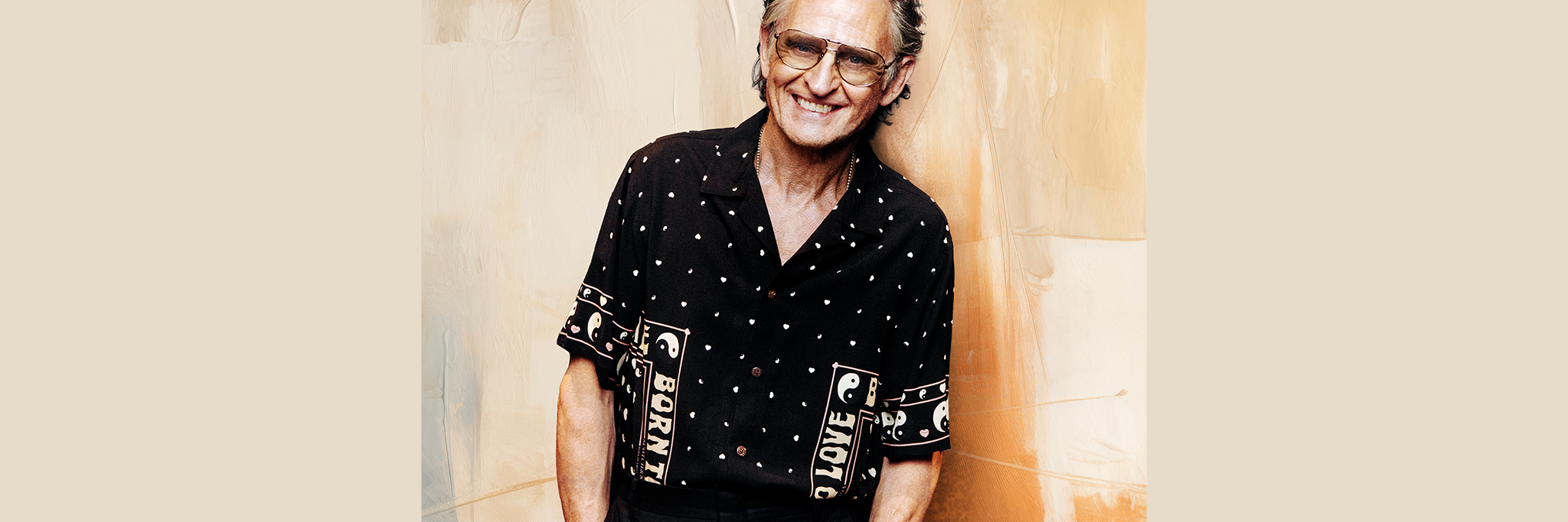 En äldre man med grått hår och glasögon ler när han står mot en beige och brun strukturerad vägg, klädd i en svartmönstrad kortärmad skjorta och svarta byxor.