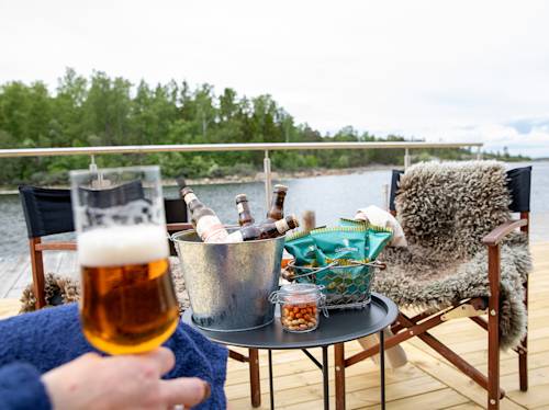 Ett bord med ölflaskor i en hink, snacks och en hand som håller ett ölglas på en terrass vid sjön med stolar och utsikt över träd och vatten.