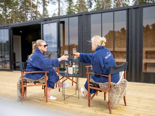 Två kvinnor i blå kläder sitter på en träterrass och skålar vid ett litet bord med drycker, utanför en modern svart byggnad omgiven av träd.