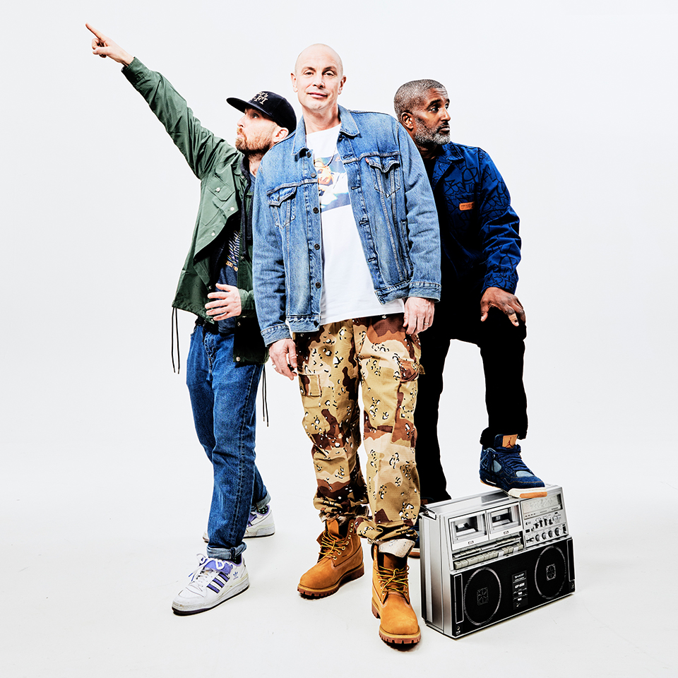 Tre män poserar självsäkert mot en vit bakgrund; en pekar uppåt, en står i mitten och en står med foten på en stor retro boombox.