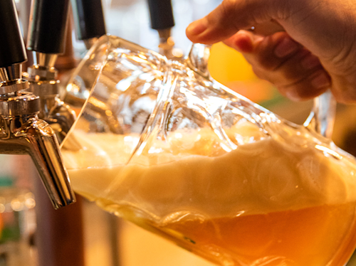 A hand tilts a glass pitcher under a beer tap as golden beer pours in, creating a thick foam head.