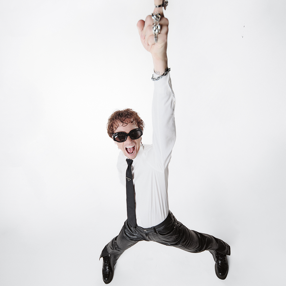 Person i solglasögon och formell klädsel står bredbent och höjer en knytnäve med kedja över huvudet mot en vit bakgrund och sträcker ut tungan.