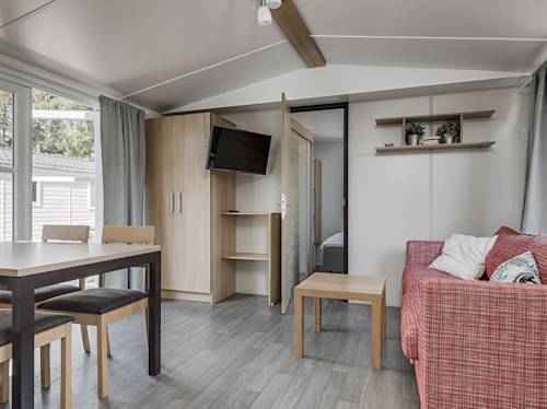 Modernt vardagsrum med röd soffa, matbord med stolar, trämöbler, vägghängd TV och utsikt mot sovrummet. Stora fönster ger naturligt ljus.
