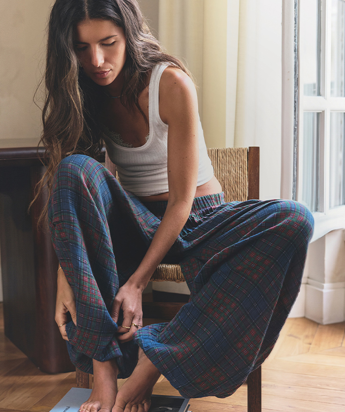 Woman sitting on wicker chair wearing blue plaid pajama pants and white tank top by sunny window on hardwood floor.