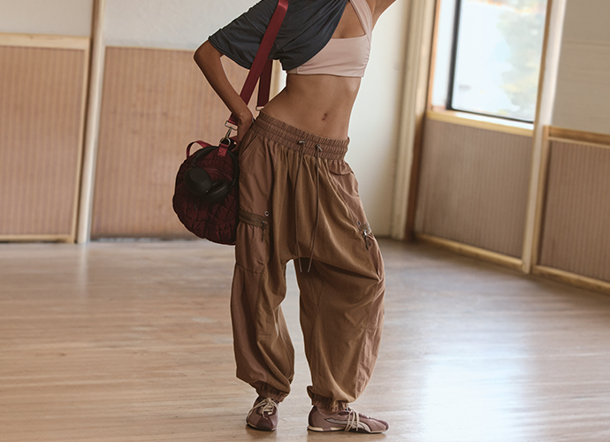 Woman in beige sports bra and brown cargo pants holding a red gym bag in a bright studio with wooden floors.