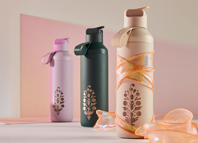 Three reusable water bottles in pink, green, and beige with tree logos and carrying straps, displayed with orange ribbon.