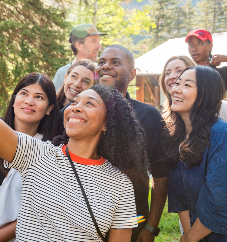 Diverse group of smiling people taking a selfie outdoors with trees and a cabin in the background.