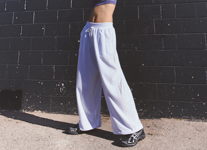 Person wearing light purple wide-leg sweatpants and black sneakers standing against a dark brick wall in sunlight.