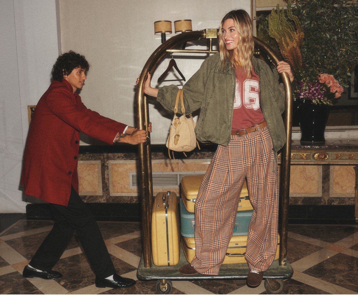 Woman in olive jacket and plaid pants stands by luggage cart with bellhop in hotel lobby. Text reads "Jackets too good to take off"