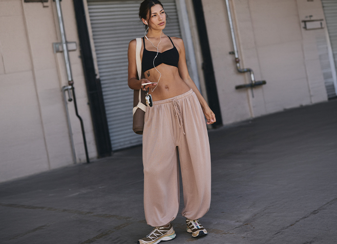 Woman in black crop top and beige wide-leg pants holding sunglasses, standing outdoors near industrial building.