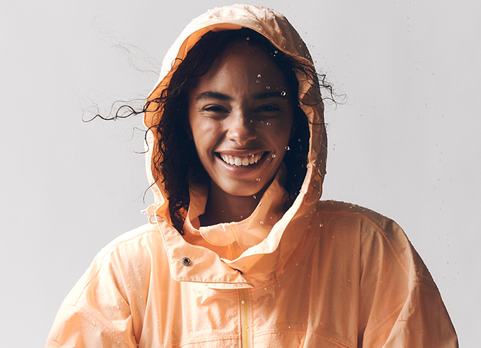 Smiling person wearing an orange hooded rain jacket with water droplets, curly hair visible, against a light background.