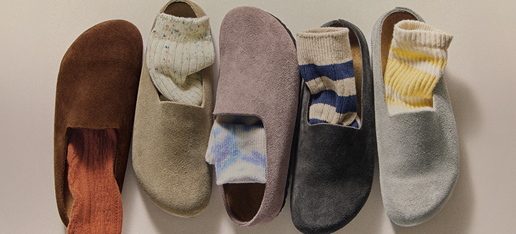 Five people wearing suede slip-on clogs in various earth tones paired with colorful socks and different pant styles.