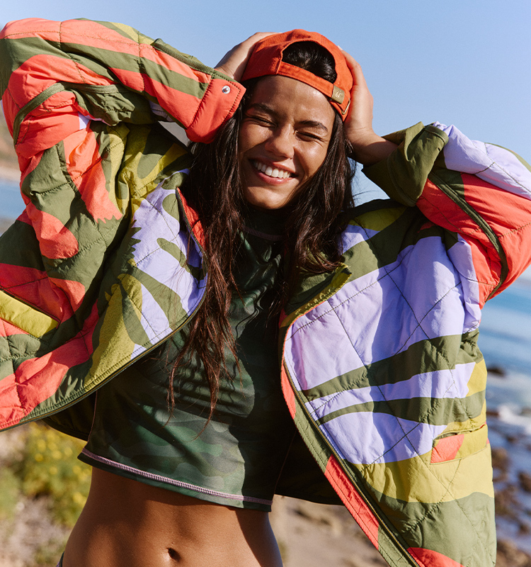Smiling woman wearing an orange cap backwards and a colorful puffer jacket in coral, green, and purple against a blue sky.