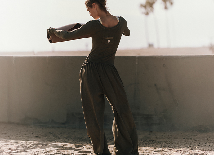 Woman in black outfit dancing on rooftop with arms extended, palm trees visible in background against pale sky.